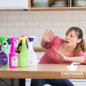A shocked woman in a pink shirt leans away from a variety of brightly colored cleaning bottles on a kitchen counter.
