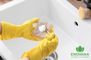 Baking soda being applied to a kitchen sponge with yellow gloves, representing Enovana Green Cleaning’s preferred eco-friendly abrasive cleaner over Bon Ami and Comet.