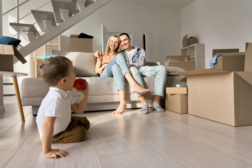 A young family surrounded by moving boxes in a bright, modern home, with a child eating an apple in the foreground. The scene illustrates the transition to a new, healthier living space supported by eco-friendly house cleaning, reflecting how Raleigh-area families benefit from sustainable cleaning routines.