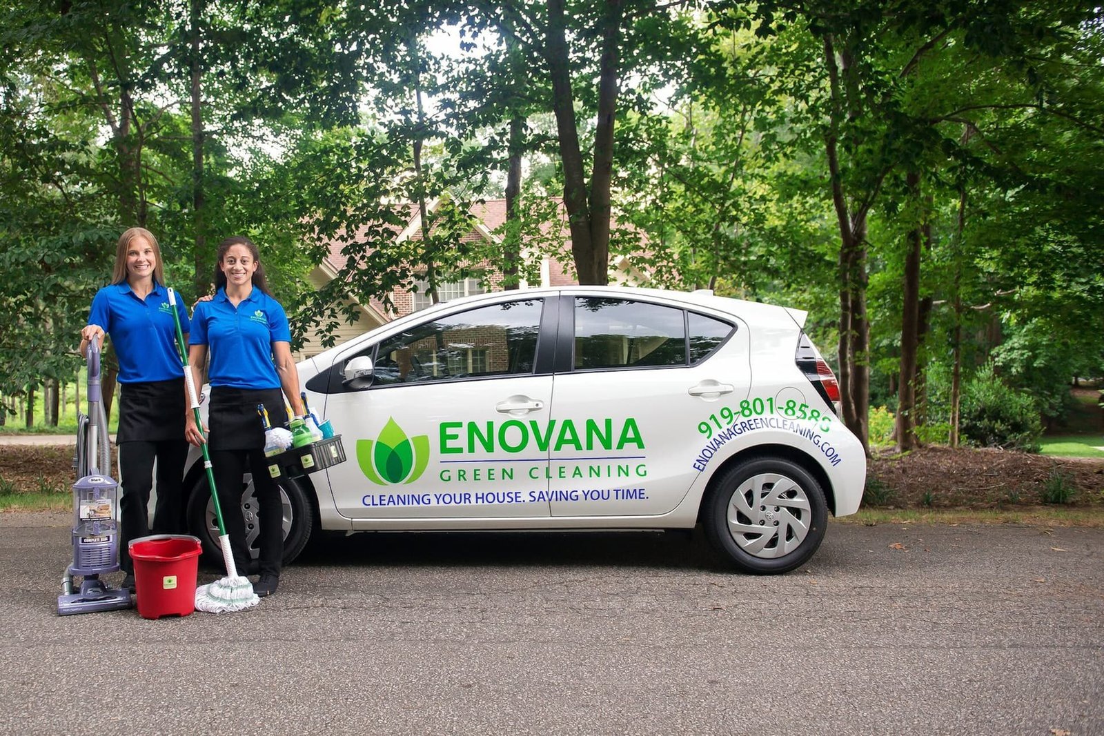 Two women wearing blue shirts and black pants stand next to a white car labeled "ENOVANA GREEN CLEANING" with cleaning supplies and equipment on a tree-lined street.